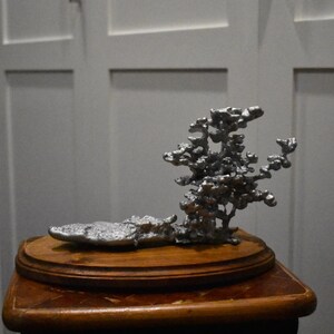 May include: A silver-colored metal sculpture with a porous, organic, and branching structure attached to a flat, elongated base. The sculpture is mounted on a smooth, oval-shaped wooden pedestal with a polished finish and visible wood grain.