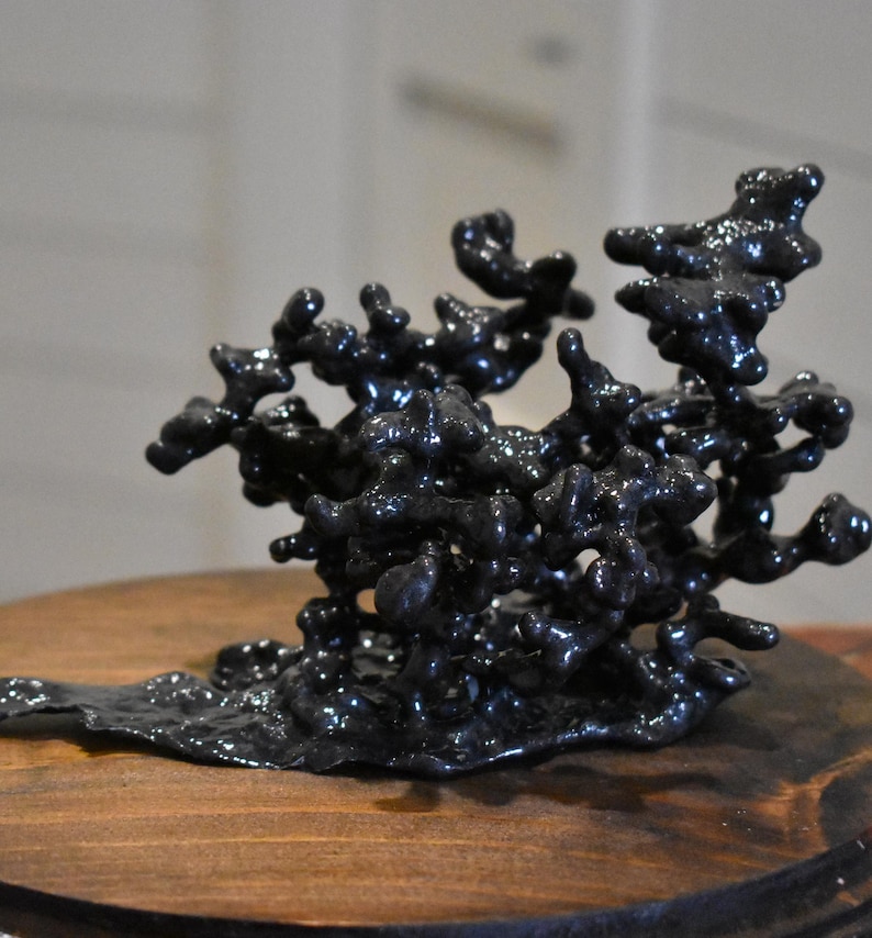 May include: A dark, glossy, black sculpture with an irregular, organic, and porous structure. The object features numerous interconnected, rounded, and bulbous protrusions that create a complex, branching, and three-dimensional form.