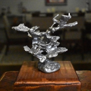 May include: A silver-colored metal sculpture with an irregular, organic, and porous structure resembling coral or molten rock. The sculpture is mounted on a square, polished wooden base with a visible grain, providing a stable foundation.
