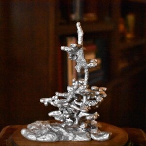 May include: A silver-colored metal sculpture featuring an organic, irregular, and porous structure with a rough, textured surface. The object consists of interconnected, molten-like branches and nodes that extend upward from a solid, uneven base.