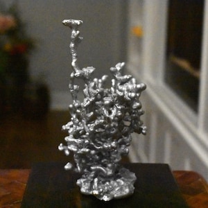 May include: A silver-colored metal sculpture with an irregular, organic, and porous structure. The object features a complex, branching, and bubbly surface texture that rises vertically from a solid, rounded base, creating a dense, abstract form.