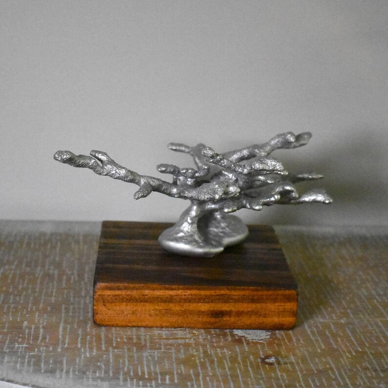 May include: A silver-toned metal sculpture shaped like a branching tree or coral formation, featuring a textured, organic surface. The sculpture is securely mounted onto a square, polished dark brown wooden base with visible wood grain patterns.