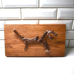 May include: A brown, rectangular wooden plaque with a bronze-colored, abstract sculpture affixed to its surface. The sculpture resembles a stylized branch or organic form. The wood grain is visible, adding texture.