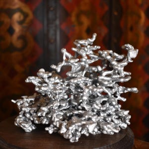 May include: A silver-colored metal sculpture featuring an intricate, organic, and branching structure with numerous rounded, interconnected nodes and textured surfaces, resembling a cast ant colony or a complex, abstract coral-like formation.