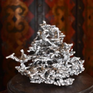 May include: A silver-colored metal sculpture featuring an intricate, organic, and porous structure with numerous interconnected branches and nodules. The object has a reflective, textured surface with a complex, multi-layered, and irregular form.