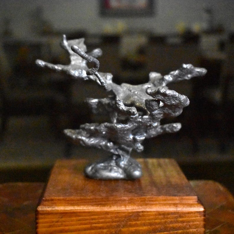May include: A silver-colored, irregular, molten-style metal sculpture with a textured, organic shape. The metallic piece is mounted on a square, polished wooden base with a visible grain, providing a stable foundation for the abstract form.