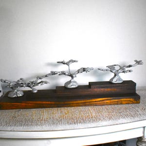 May include: A decorative display piece featuring three silver-colored, organic, branch-like metal sculptures mounted on a tiered, dark-stained wooden base. The sculptures have a textured, molten appearance and are arranged at varying heights.