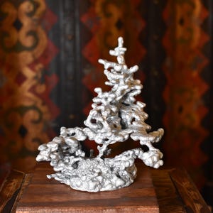 May include: A silver-colored, abstract sculpture resembling a tree. The artwork has a textured, porous surface and sits on a wooden surface. The background features a dark, patterned wall.