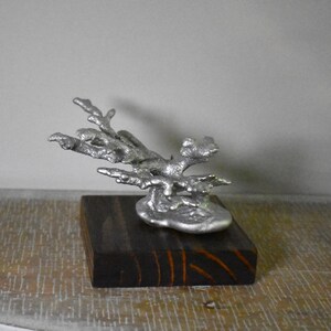 May include: A silver-colored metal sculpture shaped like a piece of coral, featuring a textured, organic form with multiple branching arms. The sculpture is securely mounted onto a square, dark brown wooden base with visible wood grain patterns.