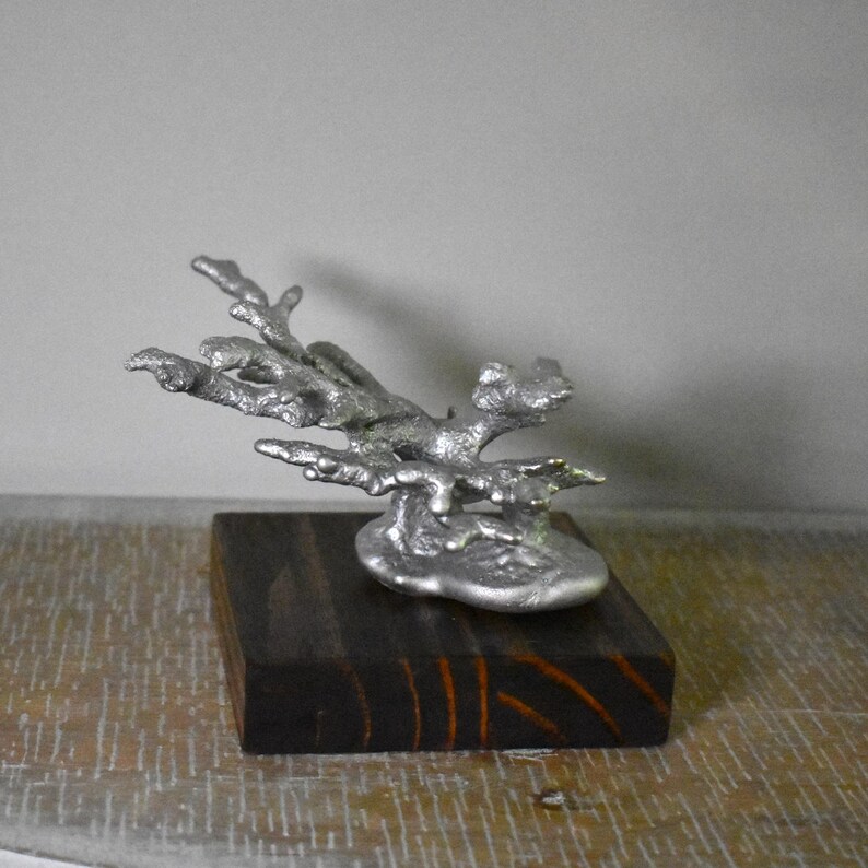 May include: A silver-colored metal sculpture shaped like a piece of coral, featuring a textured, organic form with multiple branching arms. The sculpture is securely mounted onto a square, dark brown wooden base with visible wood grain patterns.