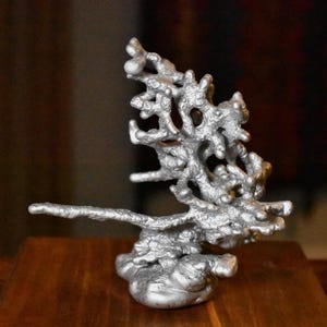 May include: A silver-colored metallic sculpture with a porous, organic, and branching structure. The object features a solid, rounded base that transitions into an intricate, upward-reaching network of textured, irregular, and interconnected protrusions.