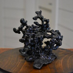 May include: A dark charcoal gray sculpture with a glossy, organic, and porous texture. The object features a complex, branching, and interconnected structure with multiple rounded protrusions and open voids throughout its irregular, three-dimensional form.