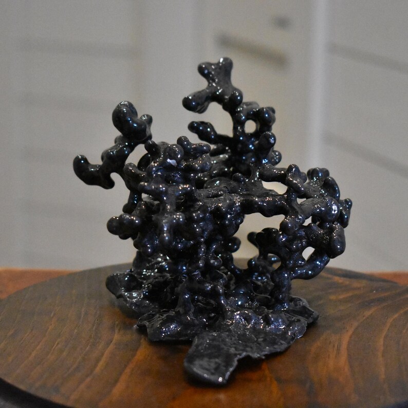 May include: A dark charcoal gray sculpture with a glossy, organic, and porous texture. The object features a complex, branching, and interconnected structure with multiple rounded protrusions and open voids throughout its irregular, three-dimensional form.