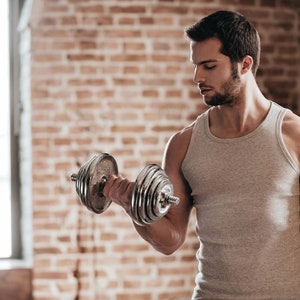 Peut inclure: Un homme portant un débardeur gris soulève une haltère argentée avec une expression concentrée. Il s'entraîne dans une pièce avec un mur en briques en arrière-plan.