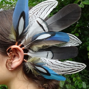 May include: A copper ear cuff with a feather design. The feathers are blue, white, and black with a striped pattern. The ear cuff is worn on the left ear.