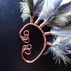 May include: A copper wire ear cuff with a decorative design and multiple feathers attached. The feathers are white, gray, and blue.