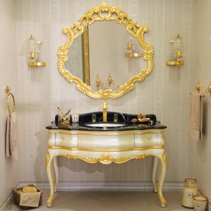 May include: A gold vanity with a black marble countertop and a large ornate gold mirror above. The vanity has a single drawer and two legs on each side. The mirror has a decorative frame with intricate details.