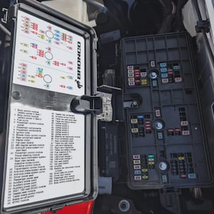 May include: An open black fuse box with a detailed diagram and a second black fuse box are visible. The diagram lists various automotive components, including the body control module, fuel pump, and starter motor. The word "BRONCO" is printed on the diagram.