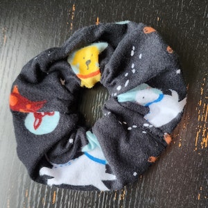 May include: Black scrunchie with a repeating pattern of cartoon dogs in yellow, white, and orange. The dogs have blue collars and are set against a background of white dots. A fun accessory for any hairstyle.
