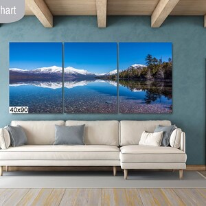 May include: A three-panel canvas print of a mountain lake with snow-capped mountains reflected in the water. The print is 40 inches by 90 inches and is hanging on a wall in a living room.