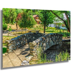 May include: A stone bridge with a curved arch spans a small stream. The bridge is made of gray and brown stones. Green trees and grass surround the bridge, with a house visible in the background.