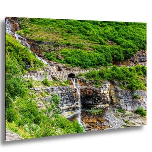 Può includere: Fotografia di un paesaggio con una cascata che scende da una scogliera rocciosa. La vegetazione verde lussureggiante ricopre il fianco della collina, con un tunnel scavato nella roccia. Una strada si snoda lungo la scogliera, aggiungendo profondità alla scena naturale.