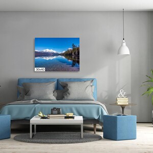 May include: A photograph of a lake with snow-capped mountains in the background. The lake is reflecting the blue sky and the mountains. The photo is framed and hanging on a wall above a bed.