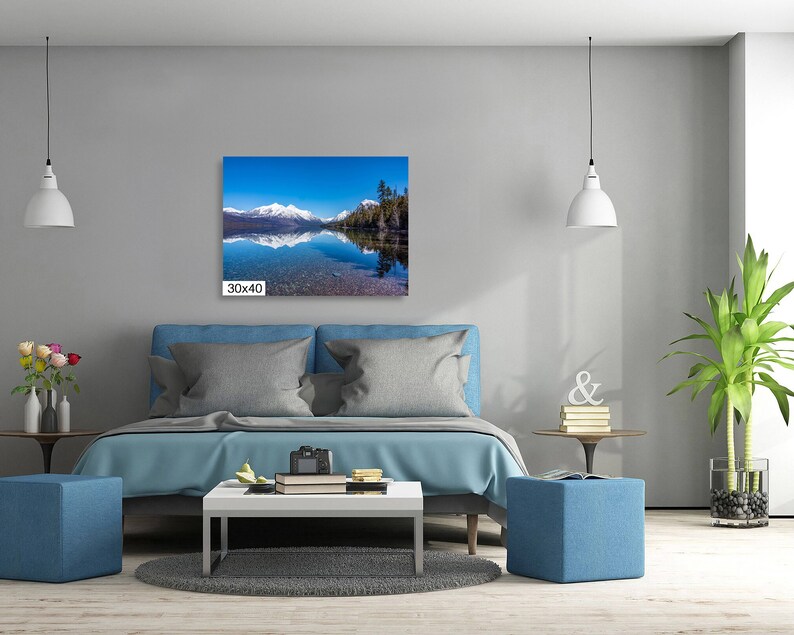 May include: A photograph of a lake with snow-capped mountains in the background. The lake is reflecting the blue sky and the mountains. The photo is framed and hanging on a wall above a bed.