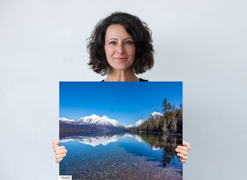 May include: A photograph of a mountain range reflected in a still lake. The mountains are snow-capped and the sky is a bright blue. The lake is a clear blue and the water is calm. The photo is printed on a 16x20 inch canvas.