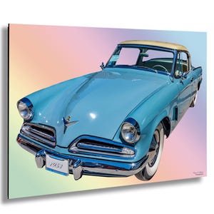 Puede incluir: Un Studebaker coupé de 1953 azul claro con detalles cromados y un interior blanco. El coche está aparcado sobre un fondo rosa pastel y azul.
