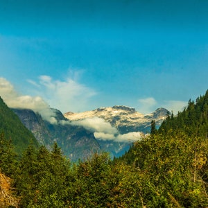 Könnte beinhalten: Blick auf eine Bergkette mit schneebedeckten Gipfeln, die teilweise von Wolken verdeckt sind. Die Berge sind mit üppigem Grün bewachsen, und der Himmel ist strahlend blau.
