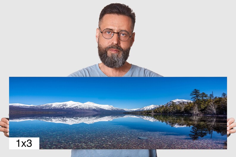 May include: A panoramic photo of a snow-capped mountain range reflected in a still lake. The sky is a bright blue, and the water is clear and calm. The photo is labeled "1x3" in the bottom left corner.