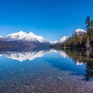 May include: A clear mountain lake with a snow-capped mountain range reflected in the water. The lake is surrounded by trees and the sky is a bright blue.