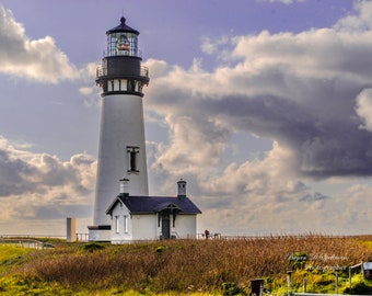 Pode incluir: Um farol branco com um telhado preto e uma lanterna de vidro no topo. O farol está situado numa colina coberta de relva com um pequeno edifício ao lado. O céu está nublado e azul.