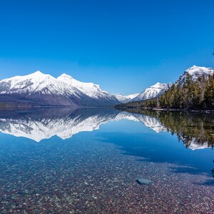 May include: A clear mountain lake with a snow-capped mountain range reflected in the water. The lake is surrounded by trees and the sky is a bright blue.