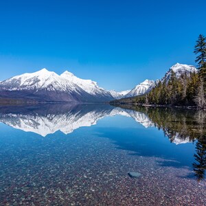 May include: A clear mountain lake with a snow-capped mountain range reflected in the water. The lake is surrounded by trees and the sky is a bright blue.