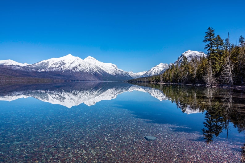 May include: A clear mountain lake with snow-capped mountains reflected in the water. The lake is surrounded by trees and the sky is a bright blue.