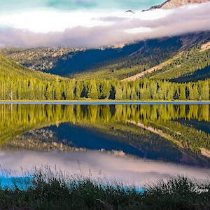 Puede incluir: Un sereno lago de montaña con un reflejo del bosque circundante y las nubes en el agua. El lago está rodeado de árboles verdes exuberantes y una cordillera en el fondo.