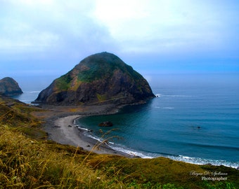May include: A scenic view of a rocky coastline with a small beach and a large, green-covered island in the distance. The sky is a light blue with some clouds.