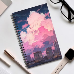 May include: A spiral bound notebook featuring a colourful illustration of a city skyline with pink and purple clouds. The notebook has a black cover.