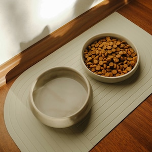 Neben dem Futternapf steht ein Wassernapf, welcher bäuchiger geformt ist und einen höheren Rand als der Futternapf hat. Der Rand ist trotzdem niedrig genug, um nicht die Schnurrhaare der Katzen zu stören.