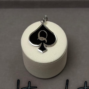 May include: A silver-toned pendant featuring a black spade design with a white 'Q' in the center, displayed on a white cylindrical stand. The pendant is set against a gray background, with the words "naughty eclectics" in cursive.