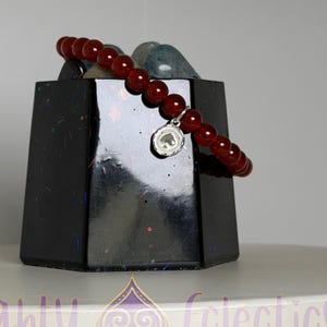 May include: A red beaded bracelet with a silver charm featuring a small design. The bracelet is displayed on a black geometric stand, with a white surface in the foreground. The text "Naughty Colectics" is visible.