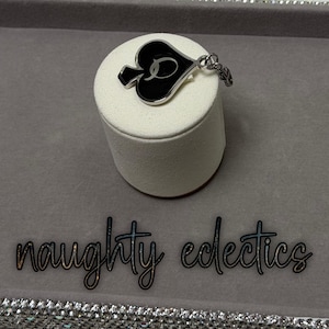 May include: A silver-toned pendant in the shape of a spade, with a black enamel center and a cursive letter "D". The pendant is on a white cylindrical display stand. The words "naughty eclectics" are below the stand.