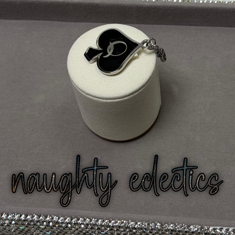 May include: A silver-toned pendant in the shape of a spade, with a black enamel center and a cursive letter "D". The pendant is on a white cylindrical display stand. The words "naughty eclectics" are below the stand.