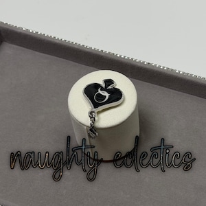 May include: A black and silver pendant in the shape of a spade with the letter "Q" inside. The pendant hangs from a silver chain and is displayed on a white cylindrical stand. The words "naughty eclectics" are below the pendant.