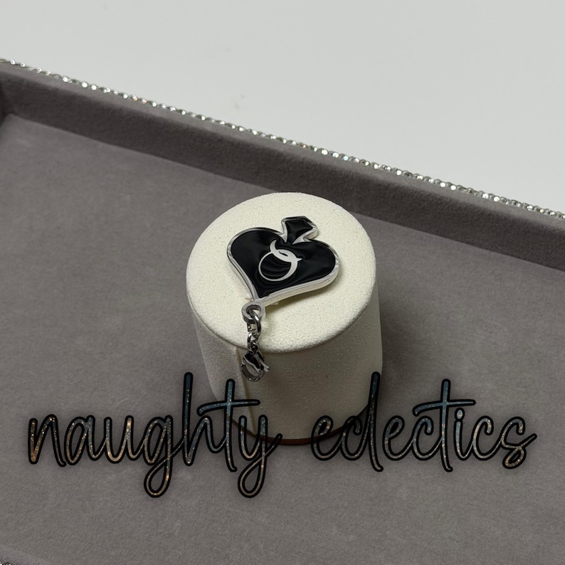May include: A black and silver pendant in the shape of a spade with the letter "Q" inside. The pendant hangs from a silver chain and is displayed on a white cylindrical stand. The words "naughty eclectics" are below the pendant.