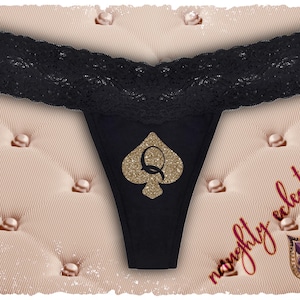 Könnte beinhalten: Schwarzer String-Tanga mit einem goldenen Glitzer-Pik-Design und dem Buchstaben "Q". Der Bund hat einen schwarzen Spitzensaum. Die Worte "naughty eclectics" sind in roter Schrift geschrieben.