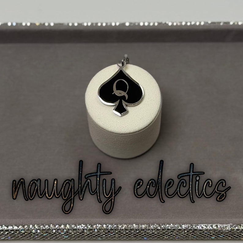 May include: A silver-toned pendant featuring a black spade design with a white 'Q' in the center, displayed on a white cylindrical stand. The pendant is set against a gray background, with the words "naughty eclectics" in cursive.