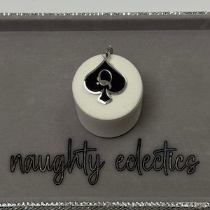 May include: A silver-toned pendant featuring a black spade design with a white 'Q' in the center. The pendant is displayed on a white cylindrical stand against a gray background. The words "naughty eclectics" are written in cursive below.